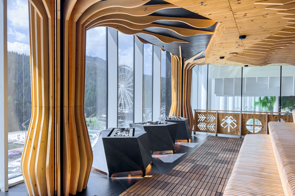 Contemporary sauna with sculptural wooden columns, faceted heaters and full height glazing with a mountain and lake view and a ferris wheel. A stylish wellness space for slow relaxation.
